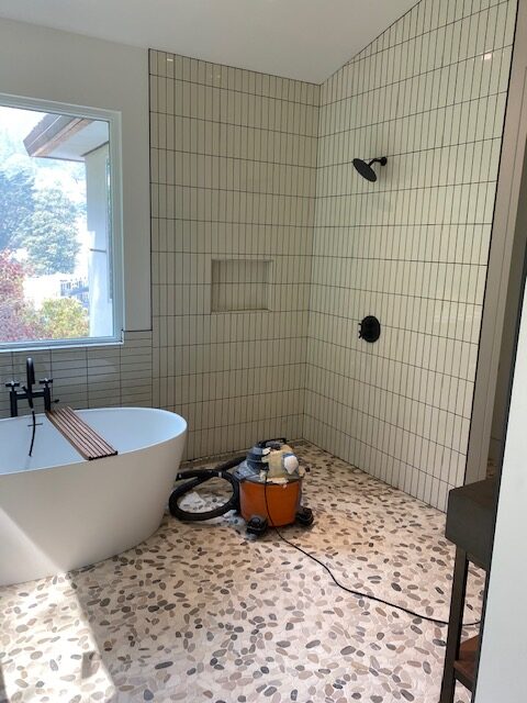 Modern bathroom featuring a freestanding tub, tiled walls, and a pebble floor. A vacuum cleaner is visible, indicating ongoing cleaning or renovation.