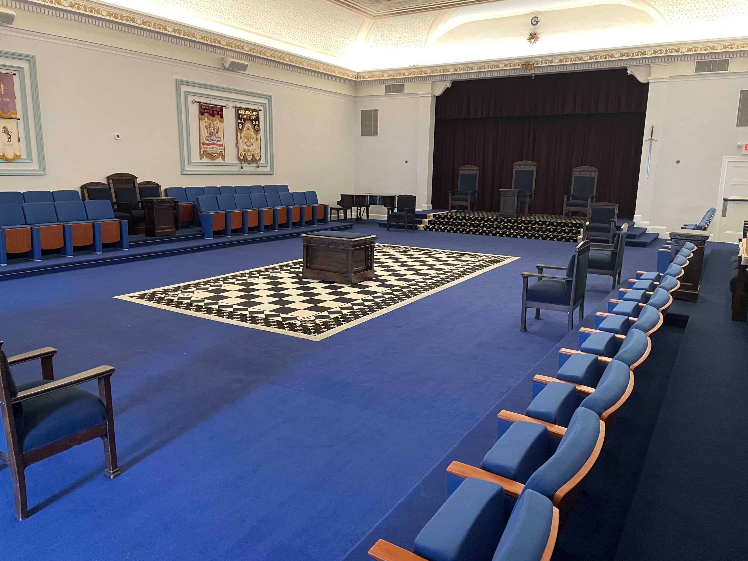 Spacious Masonic lodge interior featuring blue seating, a checkerboard floor, and seating areas for members. The setup is ready for a meeting.