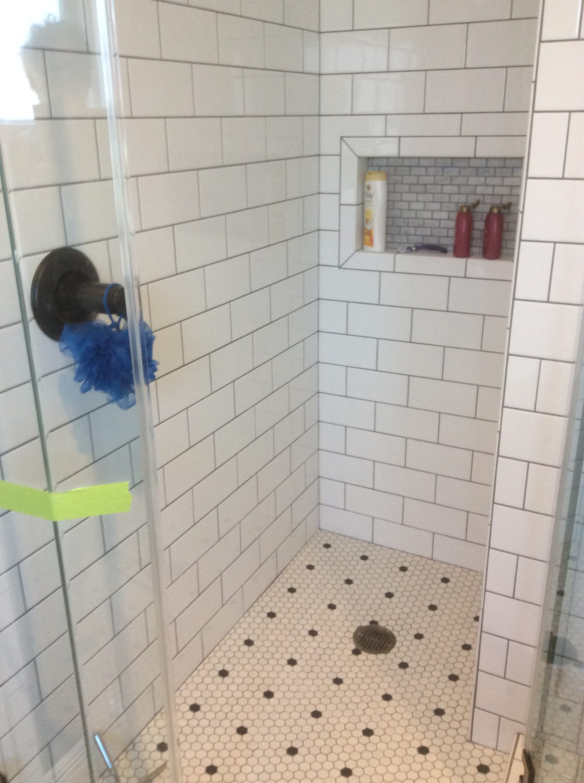 Modern shower with white subway tiles and a hexagonal black-and-white tile floor. Includes a corner shelf for toiletries and a blue loofah hanging on the glass door.