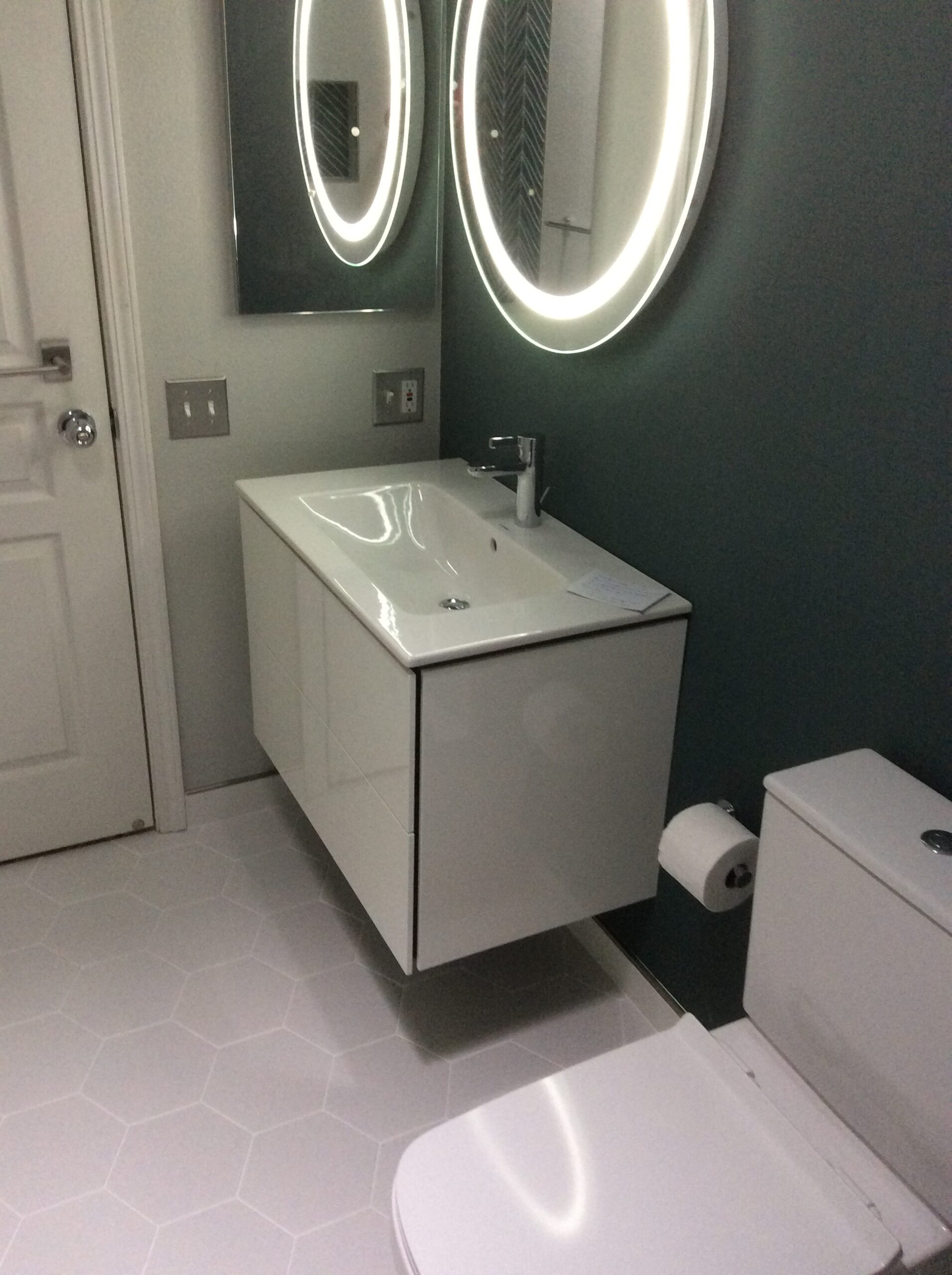 Modern bathroom featuring a white vanity with an integrated sink, illuminated round mirrors, and a minimalist design, set against a dark green wall.