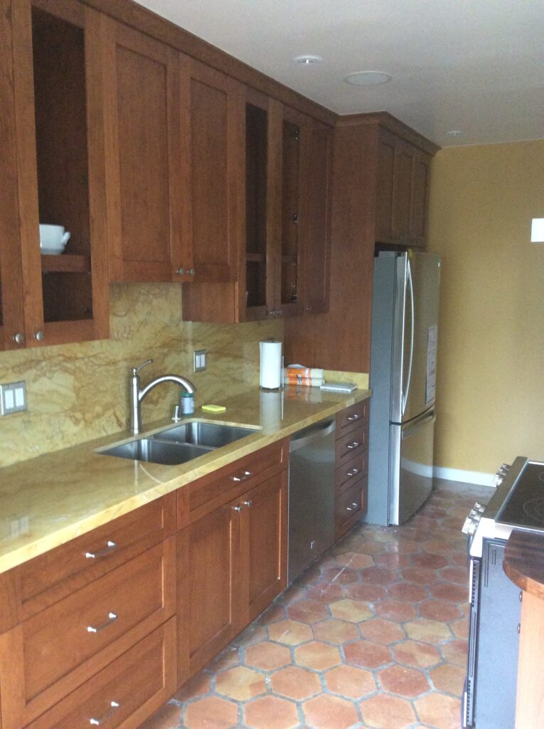 Modern kitchen featuring wooden cabinets, a stainless steel refrigerator, and a dual sink on a marble countertop, set against a terracotta tile floor.
